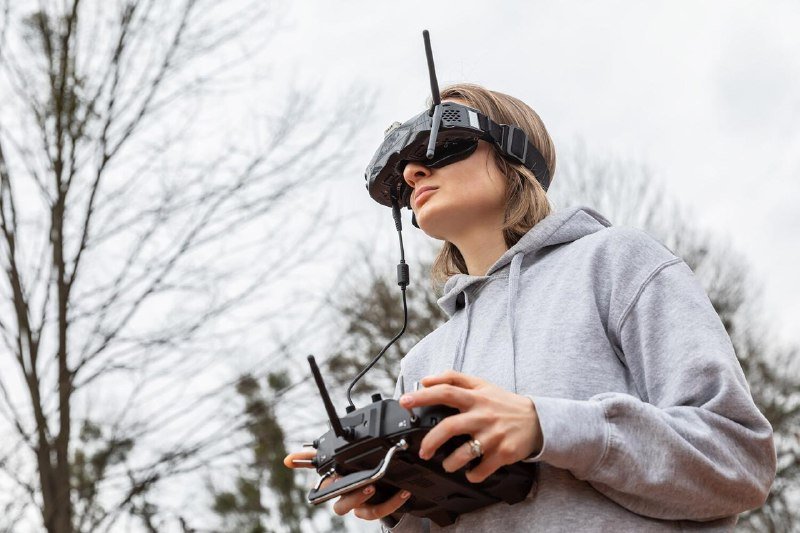A person in a gray hoodie wears FPV goggles with an antenna and holds a dual-joystick remote controller outdoors against a backdrop of bare trees.