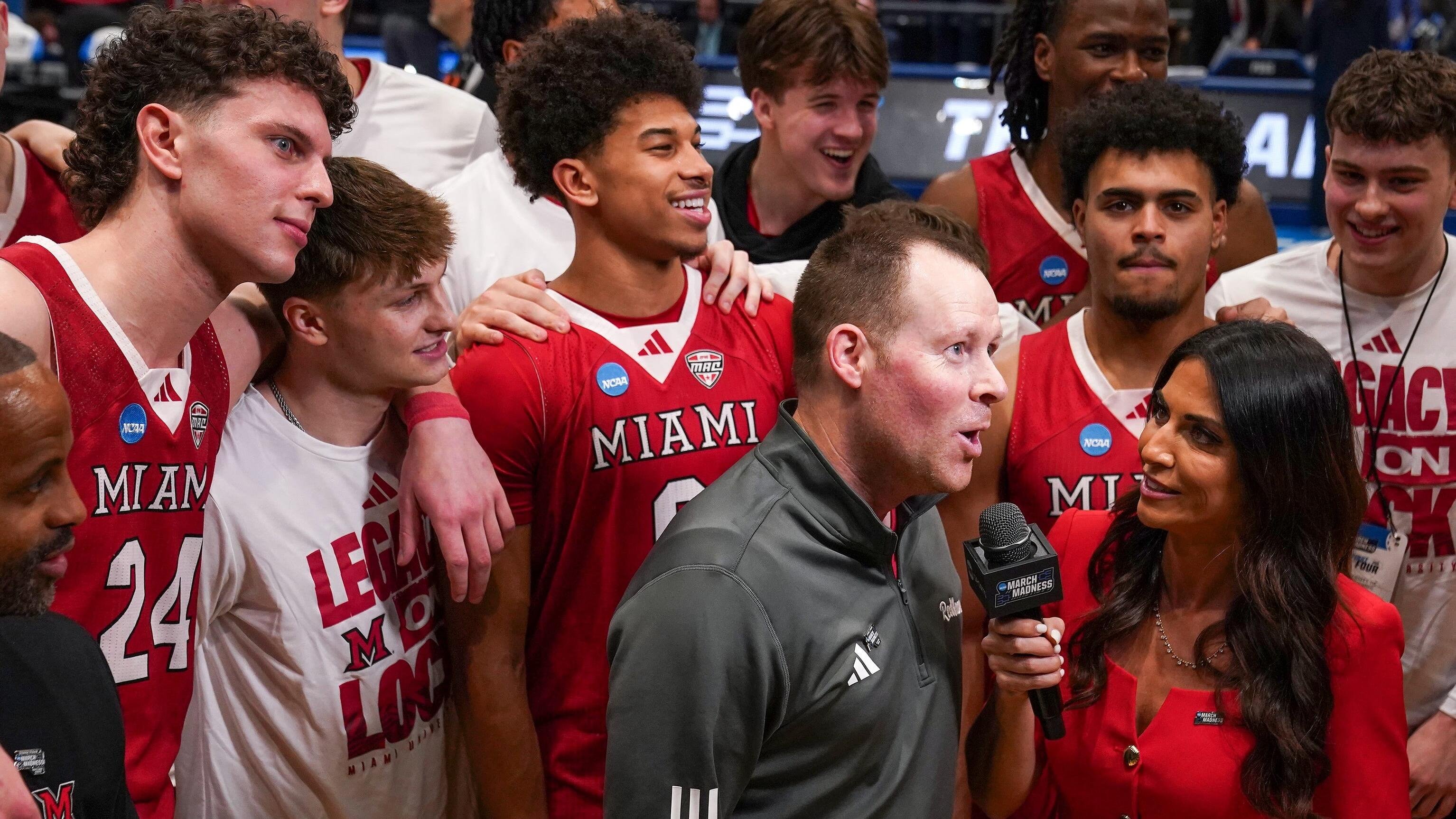 Players in red and white "Miami" jerseys celebrate with a man in a gray jacket as a woman in red interviews them courtside with a "March Madness" microphone.