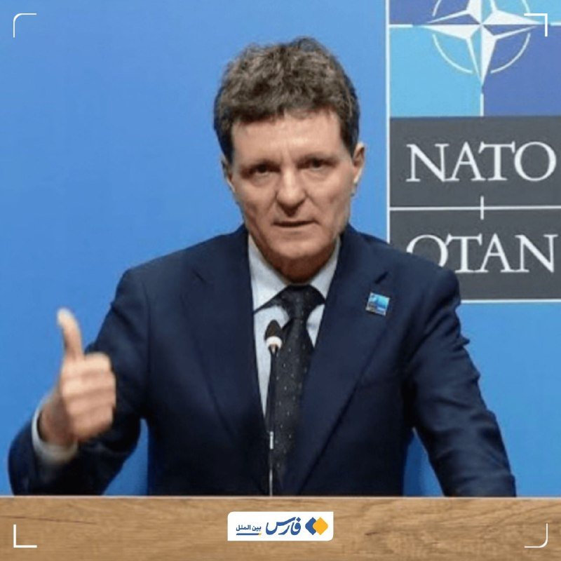 A man in a dark suit and tie speaks at a podium, gesturing with his hand, against a blue backdrop featuring the NATO logo.