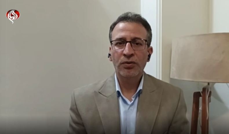 A bespectacled man in a beige blazer speaks via video call with an earpiece, seated indoors beside a lit floor lamp.
