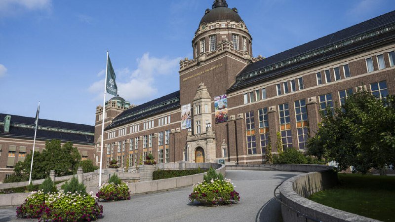 A large brick building with a prominent central dome and flagpoles stands under a blue sky, approached by a curved walkway lined with flower planters.