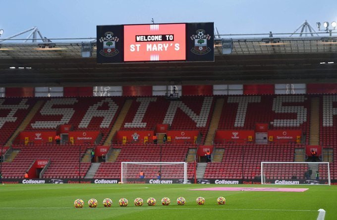 An empty football stadium displays "Welcome to St Mary's" on its digital screen, with rows of red and white seats spelling "SAINTS" behind a goalpost and footballs arranged on the pitch.