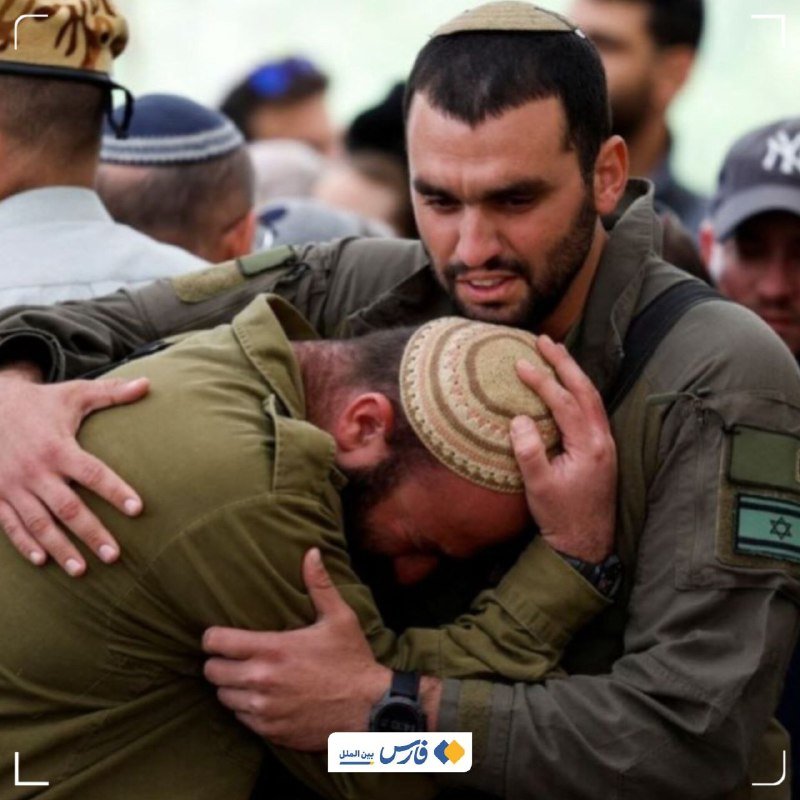 Two men in olive military uniforms embrace, one wearing a kippah, with an Israeli flag patch visible on a sleeve, surrounded by a crowd of people.