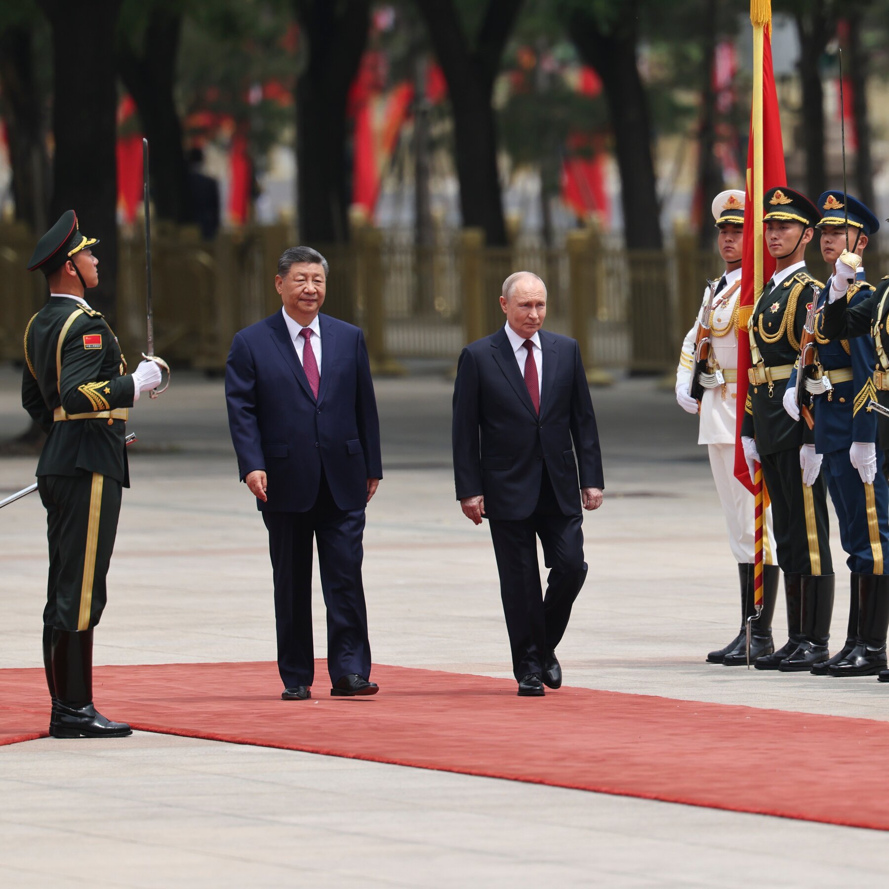 Two men in dark suits walk along a red carpet past military honor guards holding flags and swords.