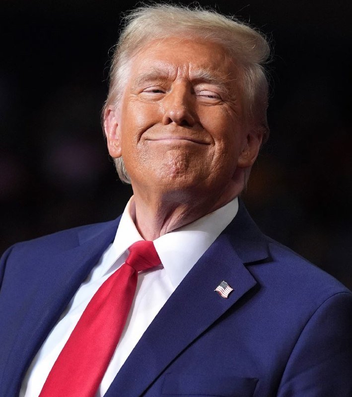 An older man with blond hair wearing a navy blue suit, white shirt, and red tie smiles with closed lips against a dark background, with a small American flag pin on his lapel.