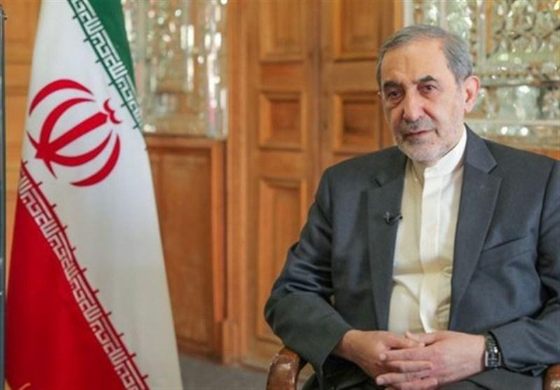 An older man with gray hair and beard, wearing a gray suit jacket over a white shirt, sits beside an Iranian flag in a room with wooden paneled walls.