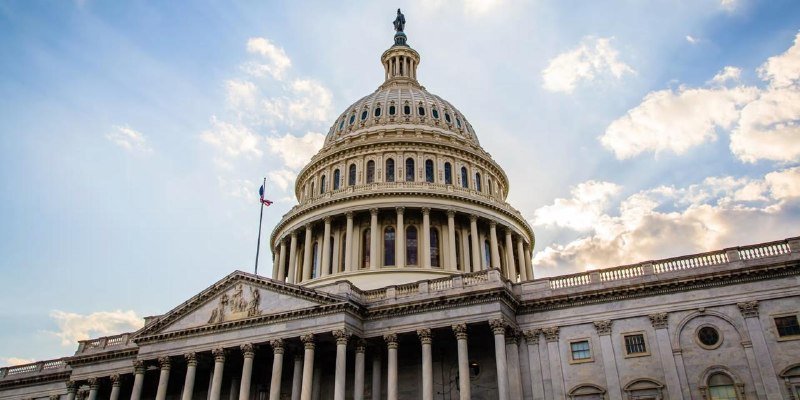 The U.S. Capitol Building's white domed structure is shown with a flag flying against a partly cloudy sky.