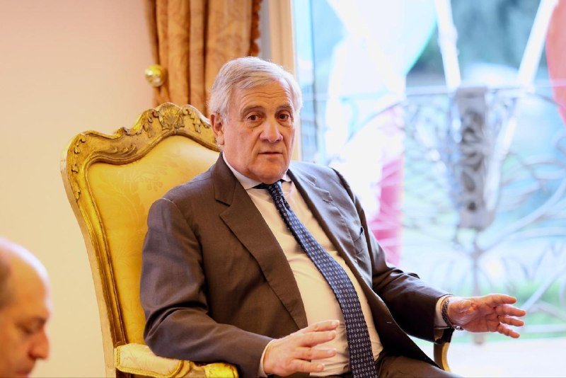 A gray-haired man in a dark suit and patterned tie gestures with both hands while seated in an ornate gold-framed chair in an elegantly furnished room.