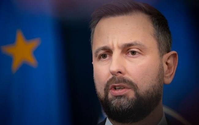 A bearded man in a suit speaks, with a blurred blue flag bearing yellow stars visible in the background.