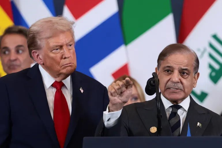 Two men in dark suits stand before a microphone, one wearing a red tie with a US flag pin, the other gesturing with a raised fist, with multiple national flags visible behind them.