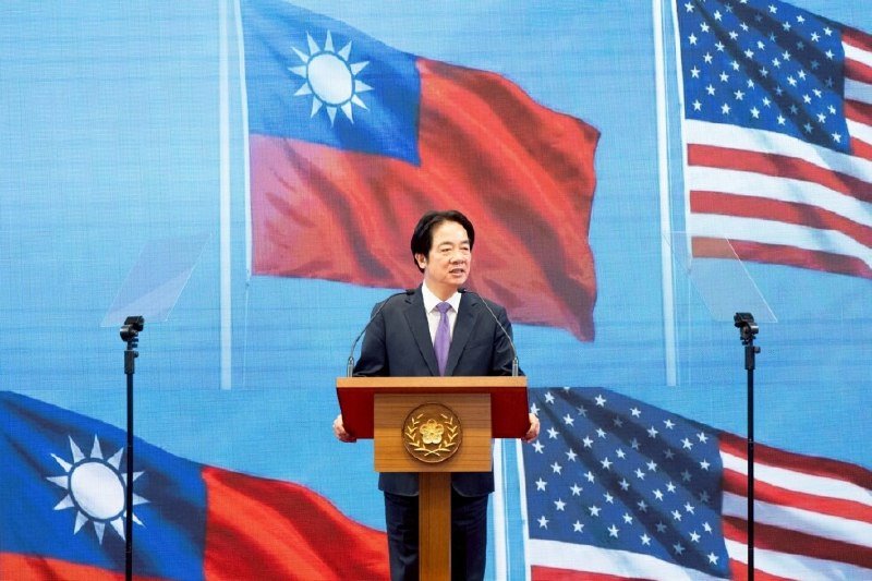 A man in a dark suit speaks at a wooden podium against a backdrop displaying Taiwanese and American flags.