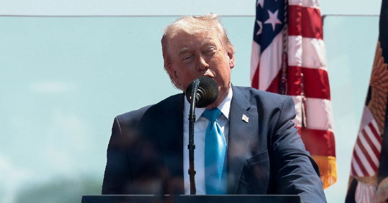 A man in a dark suit and blue tie speaks at a microphone, with an American flag visible behind him against a light backdrop.