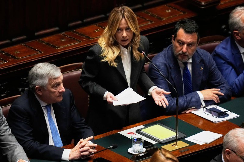 A blonde woman in a black suit speaks at a microphone while holding papers, standing beside seated men in suits at a wooden desk in a formal parliamentary chamber.
