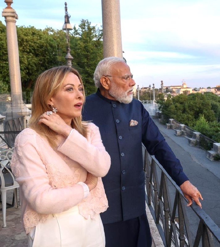 A blonde woman in a pink lace top and a gray-bearded man in a navy blue vest stand on a terrace overlooking a riverside city at dusk.