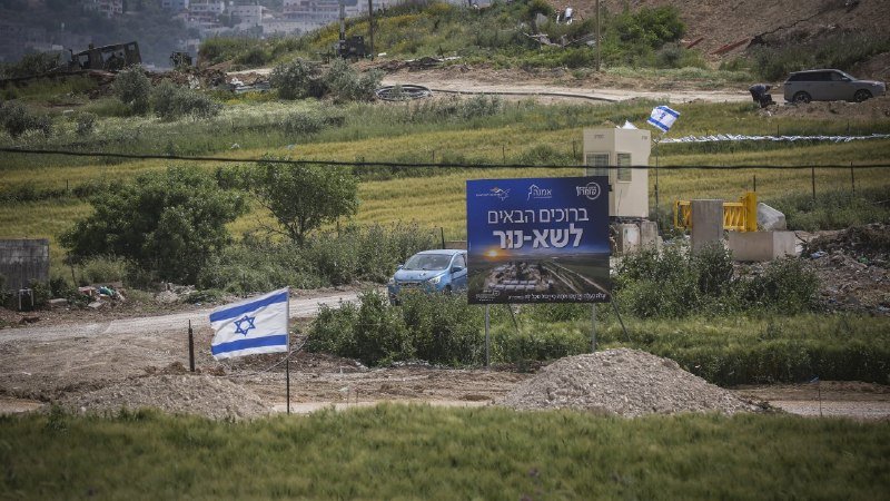 An Israeli flag flies near a Hebrew-language billboard depicting a tank, set in a rural area with parked vehicles and scattered equipment on a hillside.