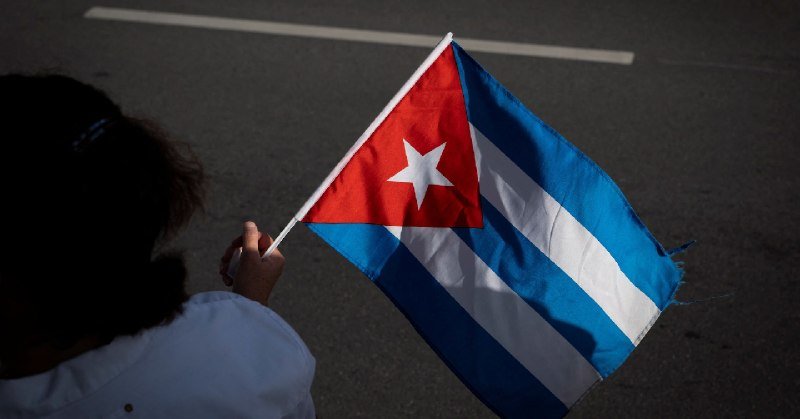 A person with dark hair holds a Cuban flag while standing on a paved street with white road markings visible.