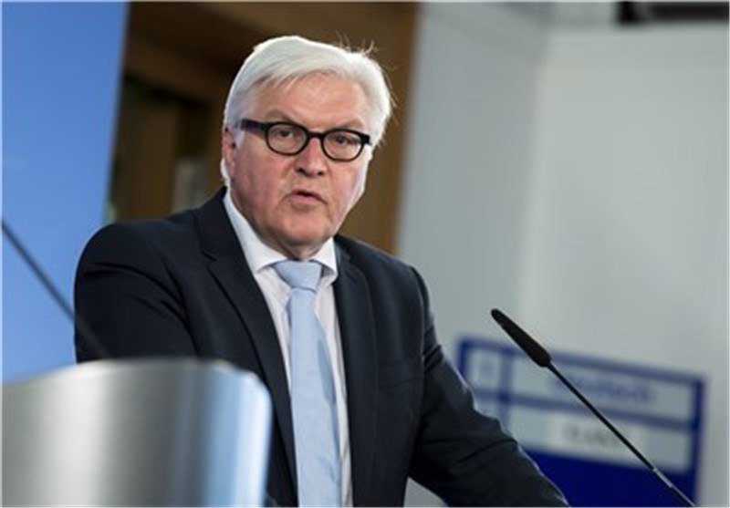 A white-haired man in a dark suit, light blue tie, and glasses stands at a podium with microphones, appearing to speak in a conference setting.