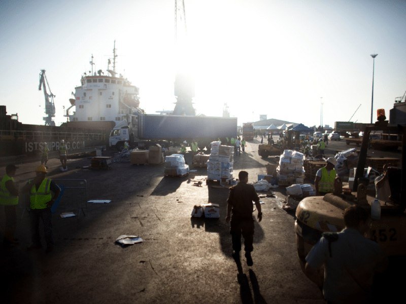 Workers in high-visibility vests unload cargo from a truck beside a docked ship at a sunlit port, with pallets and supplies scattered across the ground.