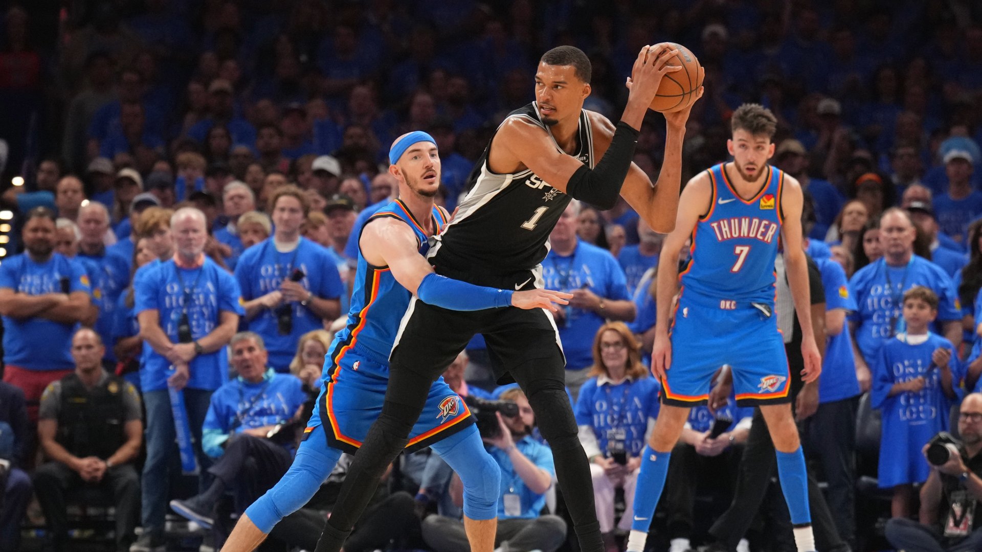 A Spurs player holds the ball up while being guarded by two Thunder players during a basketball game in front of a crowd wearing blue shirts.