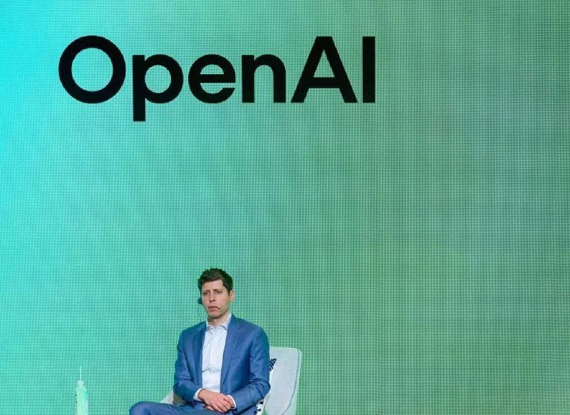 A man in a light blue blazer sits before a large green screen displaying the "OpenAI" logo.