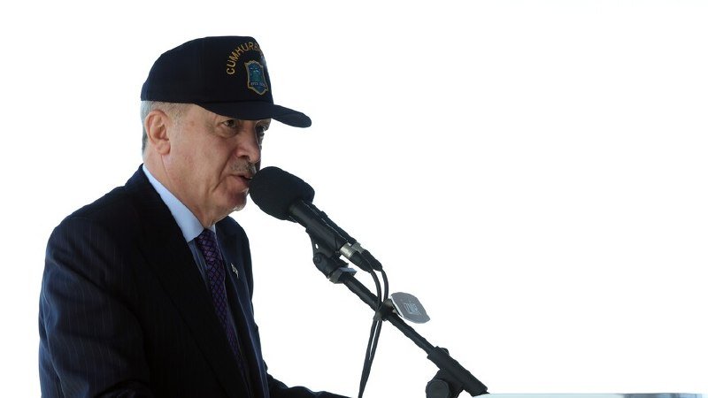 A man wearing a dark cap with "Cumhuriyet" text and a dark suit speaks into a microphone against a white background.