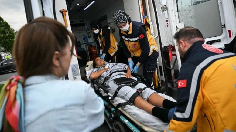 Paramedics in yellow and black uniforms with Turkish flag patches load a patient on a stretcher into the back of an ambulance.