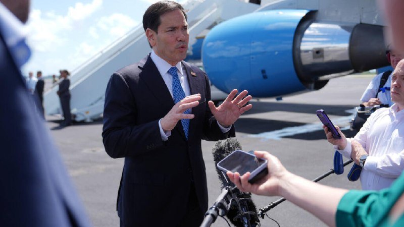 A man in a dark suit and blue tie gestures while speaking to reporters holding smartphones and microphones on a tarmac, with a blue and white airplane behind him.
