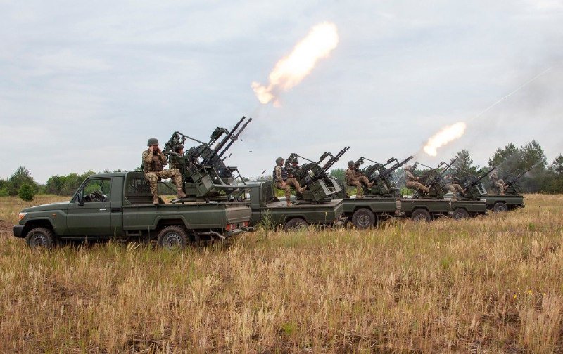 A line of green military pickup trucks mounted with anti-aircraft guns fires into a cloudy sky in a grassy field, with soldiers operating the weapons.