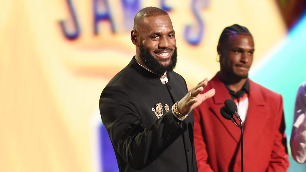 A bearded man in a black suit gestures and smiles on stage beside a man in a red suit, with a microphone and "JAMES" visible in the background.