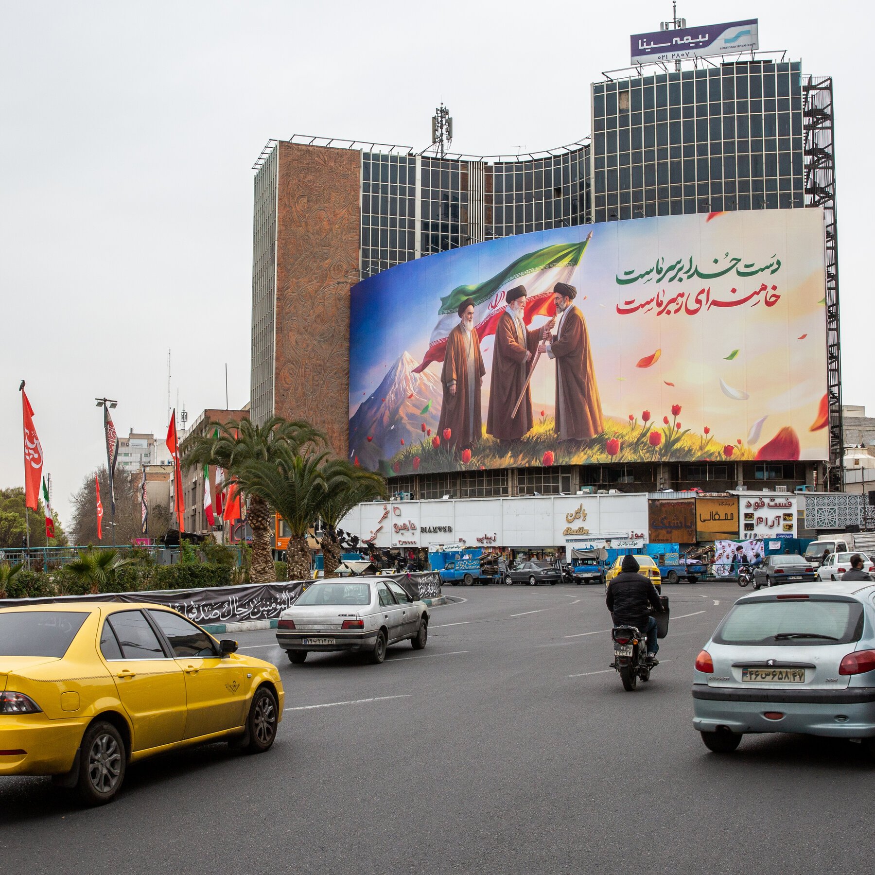 Cars and a motorcyclist travel along a city street beside a building displaying a large billboard with three robed figures, an Iranian flag, and Persian text.