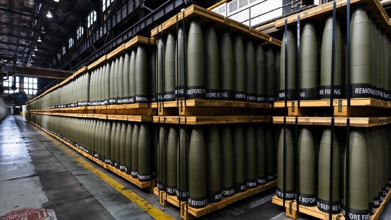 Rows of large olive-green artillery shells, labeled "REMOVE BEFORE FIRING," are stacked on wooden pallets inside an industrial warehouse.