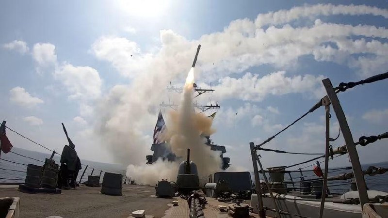 A missile launches from the deck of a naval warship at sea, with smoke billowing upward and the vessel's superstructure visible behind it.