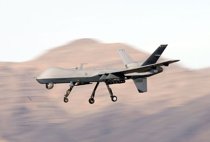 A gray military drone with landing gear deployed flies low over a blurred mountainous desert landscape.