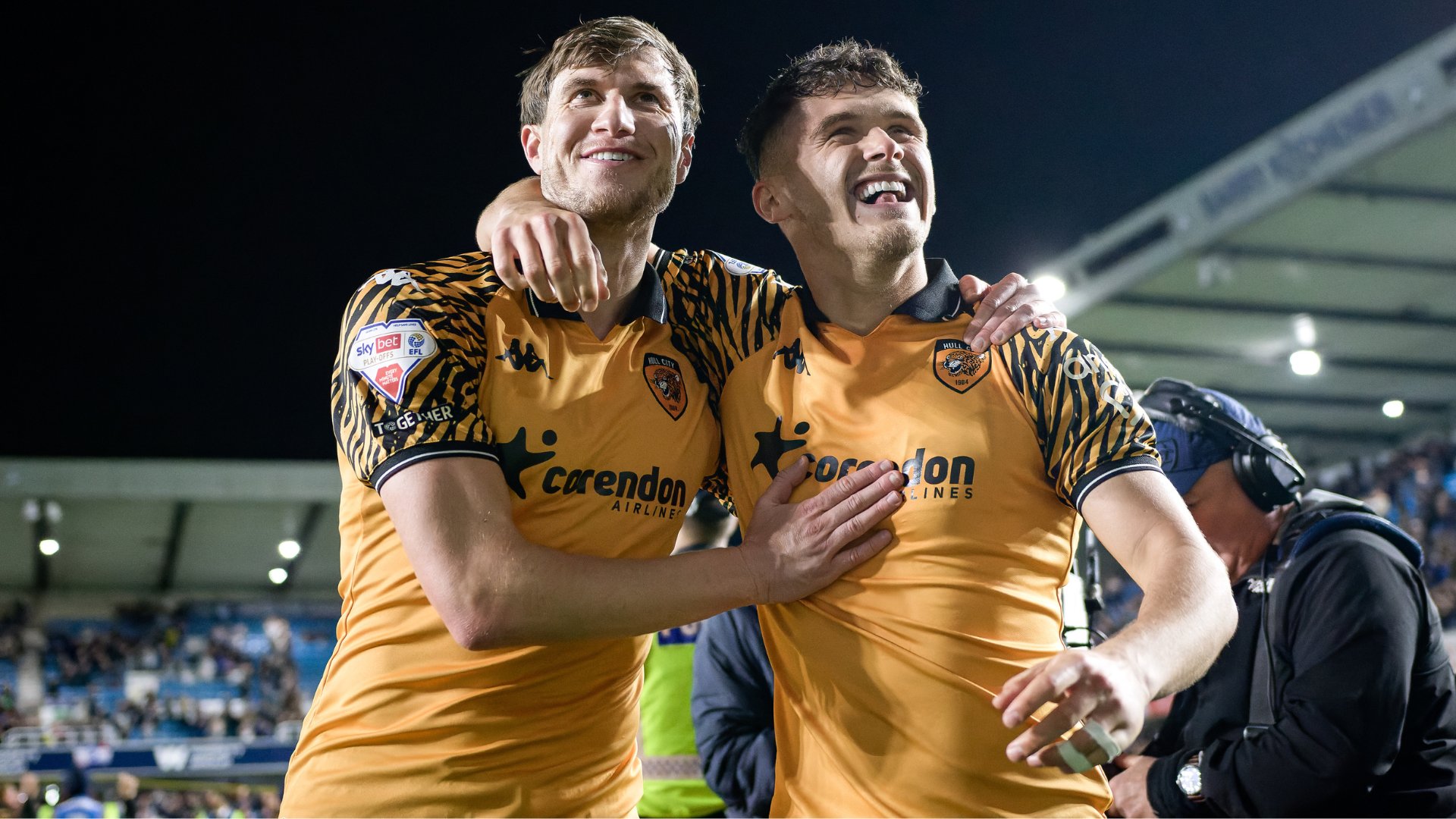 Two soccer players in orange and black tiger-striped Hull City jerseys embrace and smile in celebration on a stadium pitch at night.