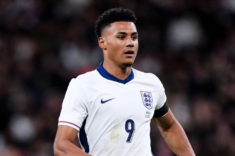 A young soccer player in a white England jersey bearing the number 9 looks on during a match, with a blurred stadium crowd behind him.