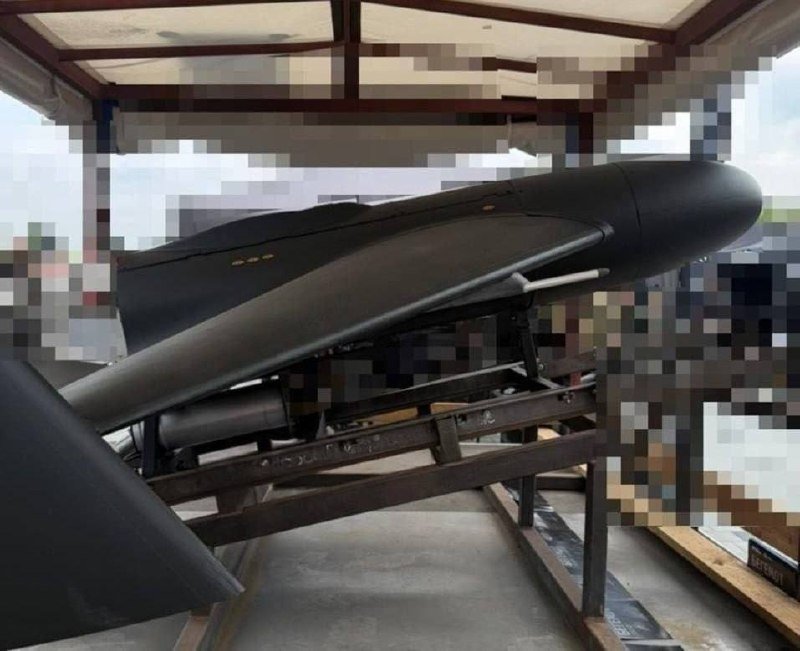 A large dark missile or torpedo rests on a metal display stand under a covered outdoor shelter, with partially pixelated signage visible in the background.