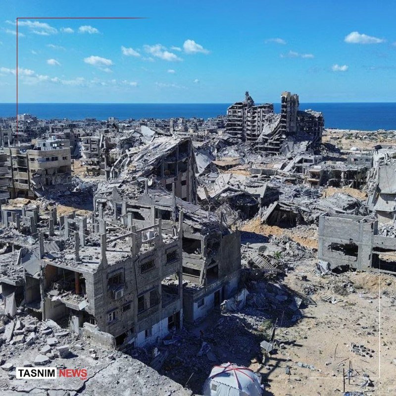 Extensive urban rubble and destroyed multi-story buildings stretch toward the sea under a clear blue sky, with a "TASNIM NEWS" watermark visible.