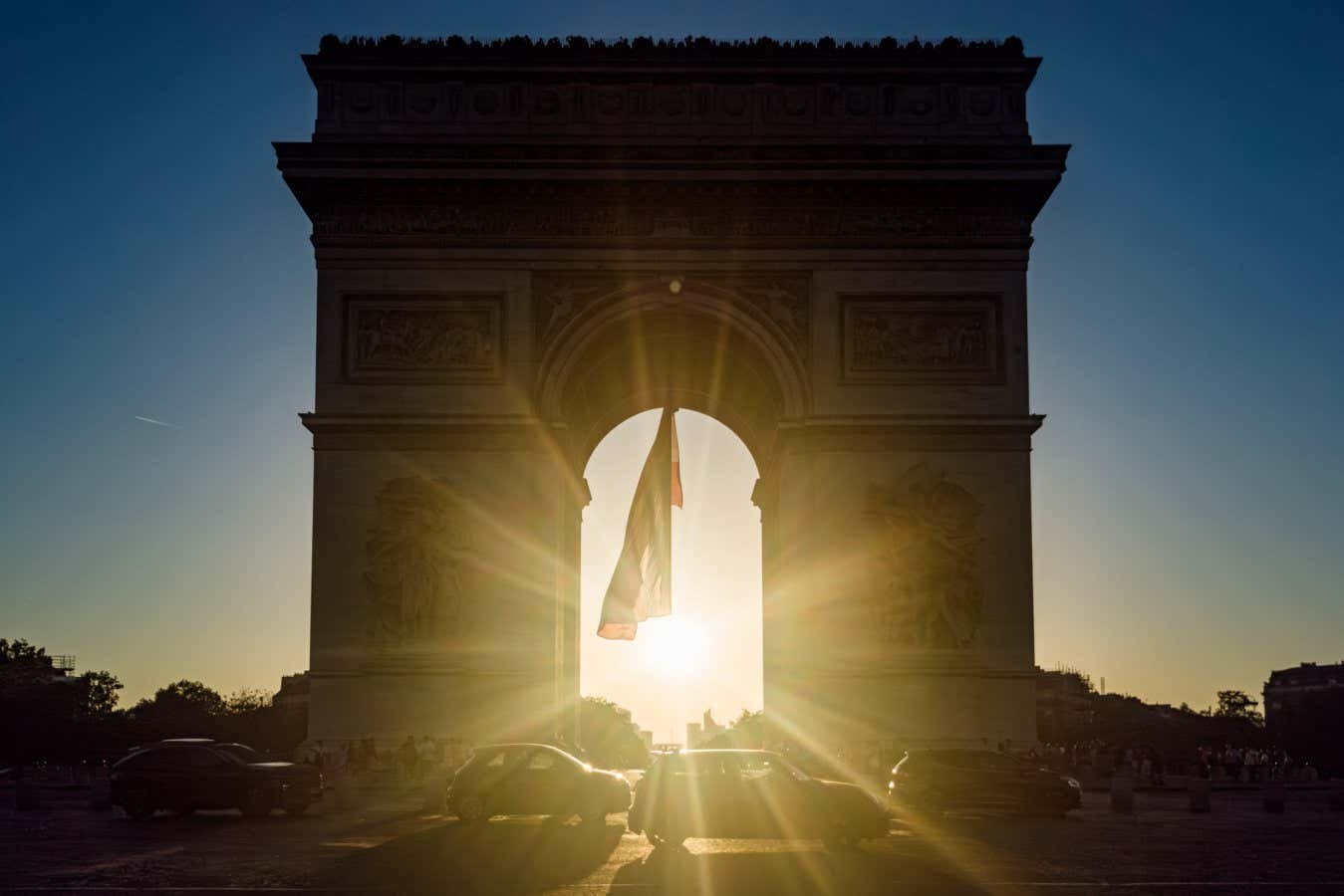 A silhouetted triumphal arch frames a sunset, with a flag and cars passing through its central opening.