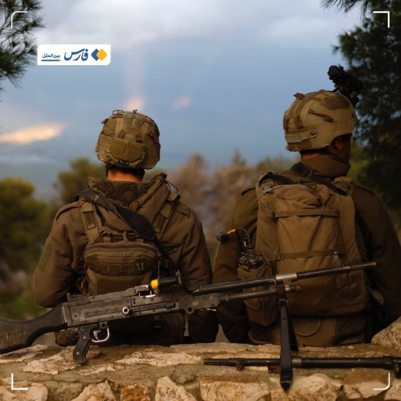 Two helmeted soldiers in combat gear with backpacks sit on a stone wall, one holding a mounted machine gun, viewed from behind against a cloudy sky.