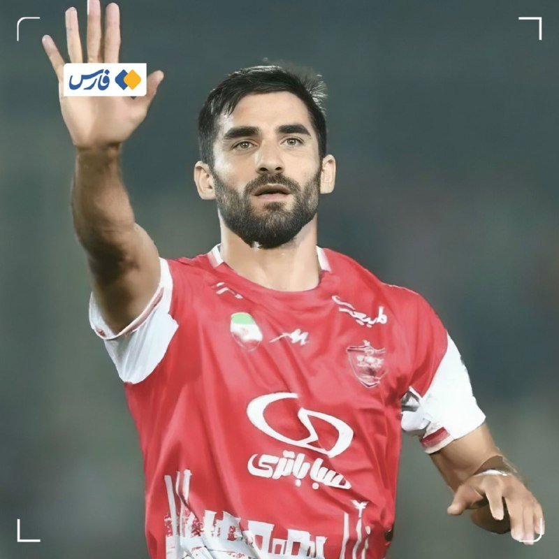 A bearded soccer player in a red and white jersey raises his hand with an open palm during a match.