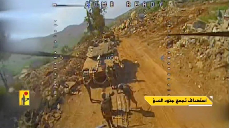 A military helicopter gunsight view shows a camouflaged tank driving along a dirt road in a rocky, mountainous area, with Arabic text on a news broadcast graphic.