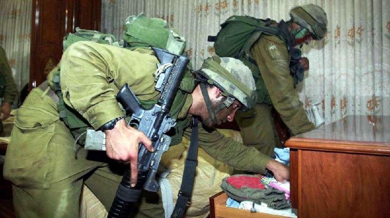 Armed soldiers in green uniforms and helmets search the contents of an open dresser drawer inside a room with floral curtains.