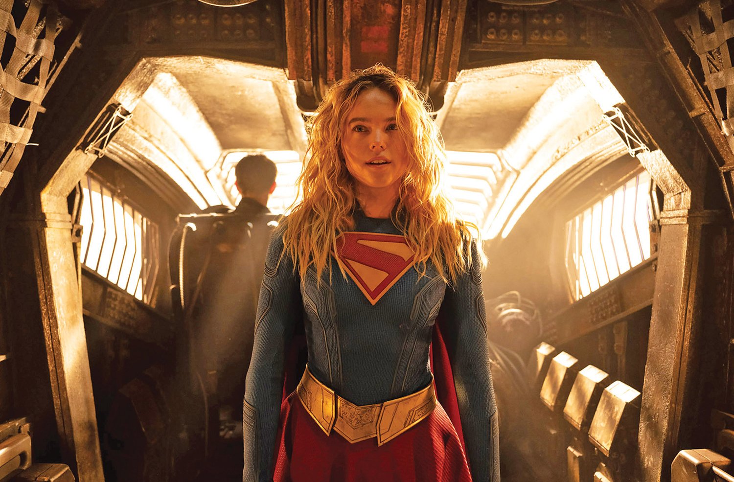 A blonde woman in a blue and red superhero costume with a diamond chest emblem stands in a dimly lit, industrial metallic corridor.
