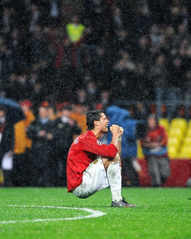 A soccer player in a red jersey and white shorts crouches on the grass field, fist raised to his mouth, with photographers and a crowded stadium visible behind him.
