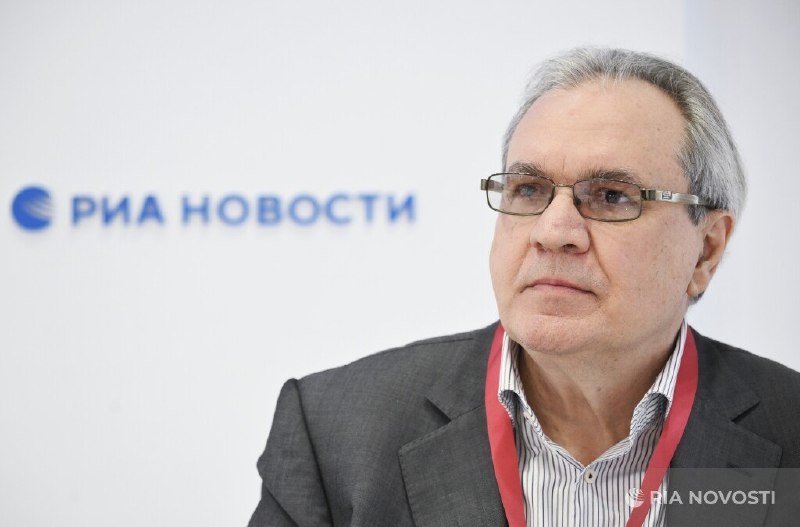 A gray-haired man in glasses and a gray blazer wearing a red lanyard looks off-camera, with a backdrop displaying the "РИА НОВОСТИ" logo.