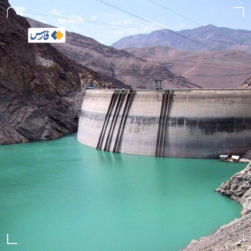 A large concrete dam holds back turquoise-green reservoir water, surrounded by arid, rocky mountains under a partly cloudy sky.