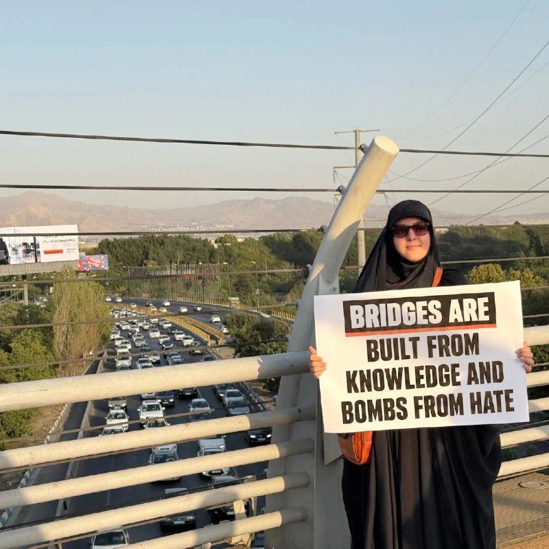 A woman in a black chador holds a sign reading "BRIDGES ARE BUILT FROM KNOWLEDGE AND BOMBS FROM HATE" on a bridge overlooking heavy traffic.