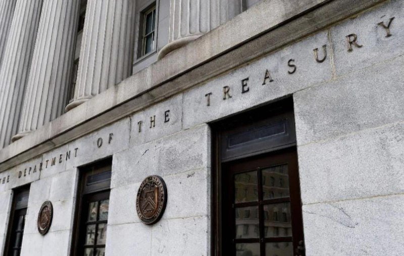The stone facade of a building displays the carved inscription "THE DEPARTMENT OF THE TREASURY" above windows and bronze seals.