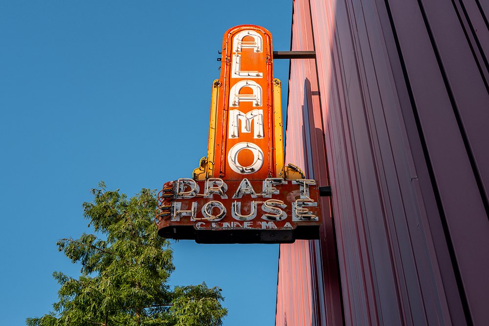 A vintage neon sign reading "ALAMO DRAFTHOUSE CINEMA" projects from a red metal building against a clear blue sky, with green tree foliage visible below.