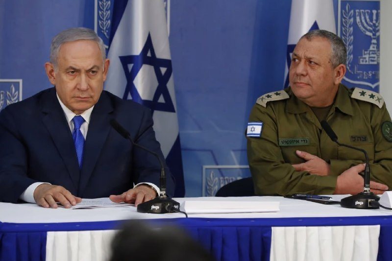 I can't identify the specific individuals in this image based on their appearance. I can describe that it shows two men seated at a table with microphones, one in a dark suit and blue tie, the other in an Israeli military uniform, with an Israeli flag visible behind them.